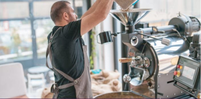 Photograph of a man wearing apron, pouring coffee beans into a roaster